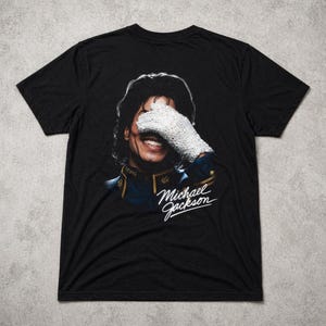 May include: Black t-shirt featuring a portrait of Michael Jackson, with his face partially covered by a white glove. The shirt includes the signature "Michael Jackson" in white script.