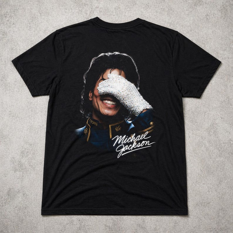 May include: Black t-shirt featuring a portrait of Michael Jackson, with his face partially covered by a white glove. The shirt includes the signature "Michael Jackson" in white script.