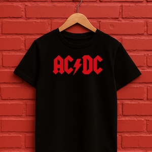 May include: Black t-shirt featuring the red AC/DC logo and lightning bolt. The garment is suspended on a wooden hanger, set against a red brick wall. The design is centrally placed on the front.