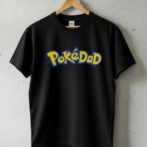 May include: Black t-shirt featuring the text "PokéDad" in yellow and blue, a parody of the Pokémon logo. The garment is suspended on a wooden hanger against a grey wall.