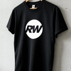 May include: Black t-shirt with a white circular graphic. The graphic features the letters "RW" and a star. The t-shirt is hanging on a wooden hanger against a grey wall. The shirt is made of a soft material.