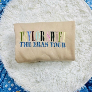 May include: A beige sweatshirt embroidered with the words "TAYLOR SWIFT THE ERAS TOUR" in multiple colours. The sweatshirt is placed on a white, fluffy, circular rug, against a blue patterned background.