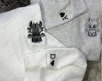 May include: White and gray sweatshirts with embroidered dragon designs. One sweatshirt features a black dragon with green eyes, while another has a gray dragon. The sleeves display a letter and a heart.