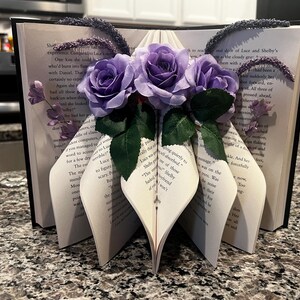 May include: An open book with pages folded into a decorative shape, adorned with purple roses and greenery. The book's pages are visible, and the spine is black. Lavender-colored accents are placed on the top of the book.