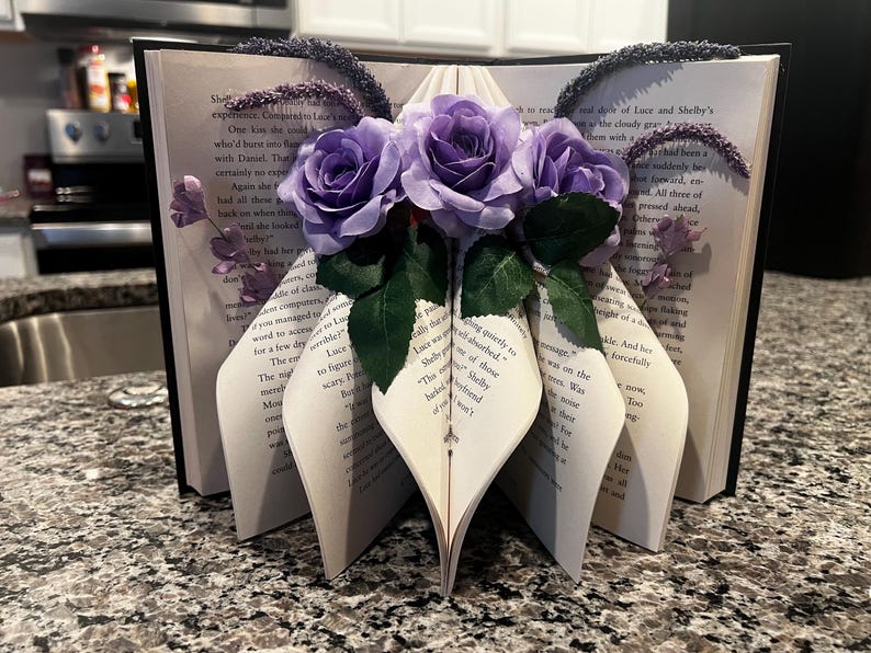 May include: An open book with pages folded into a decorative shape, adorned with purple roses and greenery. The book's pages are visible, and the spine is black. Lavender-colored accents are placed on the top of the book.
