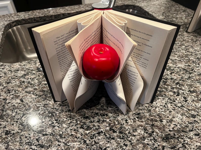 May include: A decorative book with pages folded to cradle a bright red apple. The book has a black cover and the pages are open, revealing printed text. The apple is smooth and round, adding a pop of color.