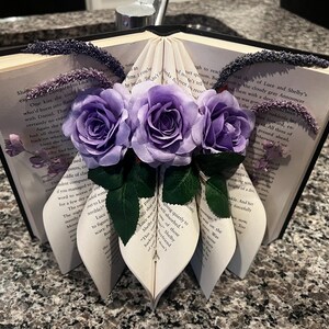 May include: A decorative book sculpture with pages folded into a heart shape, adorned with three purple roses and green leaves. The book's edges are embellished with dark purple accents, creating a unique floral arrangement.