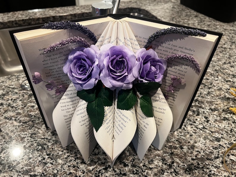 May include: A decorative book sculpture with pages folded into a heart shape, adorned with three purple roses and green leaves. The book's edges are embellished with dark purple accents, creating a unique floral arrangement.