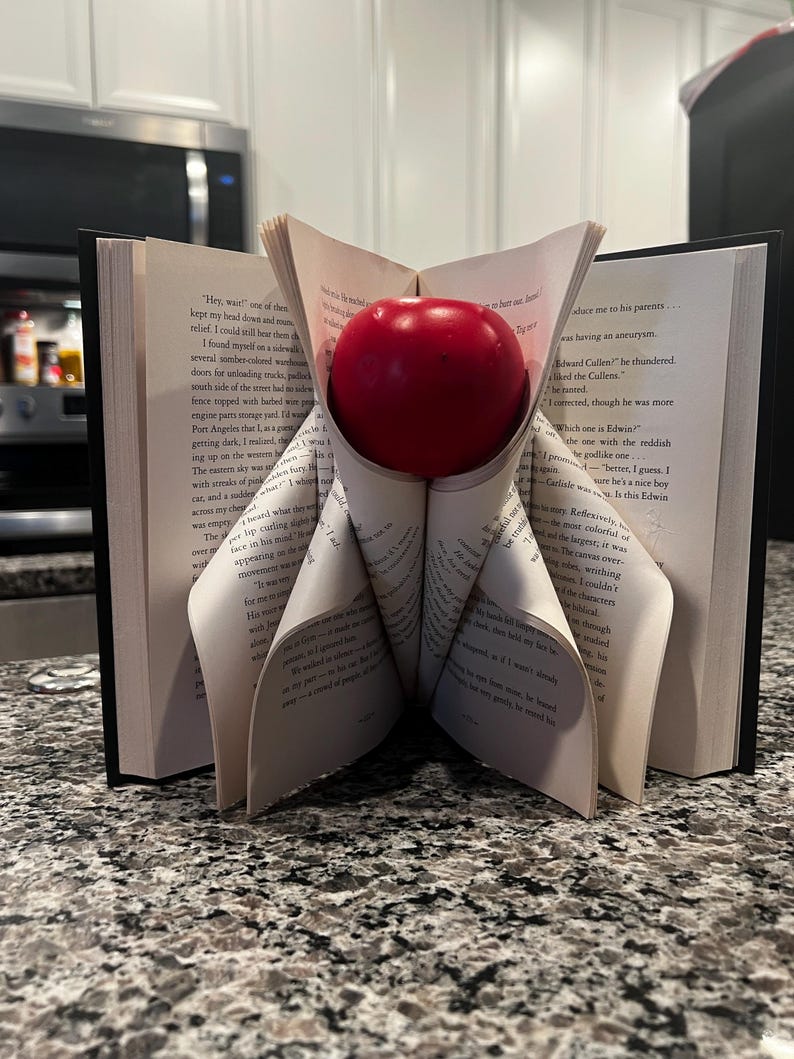 May include: A red apple rests in a book, with pages folded to create a unique display. The open book stands upright on a speckled countertop, with the apple centered. The image showcases a creative book art project.