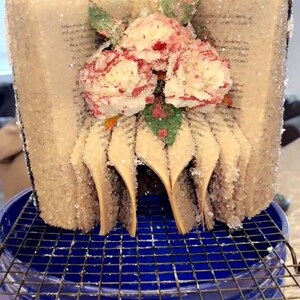 May include: A unique art piece crafted from a book, shaped into a floral arrangement. The pages are visible, with a dusting of glitter. Red and white flowers with green accents adorn the top. The piece sits on a blue bucket with a metal grate.