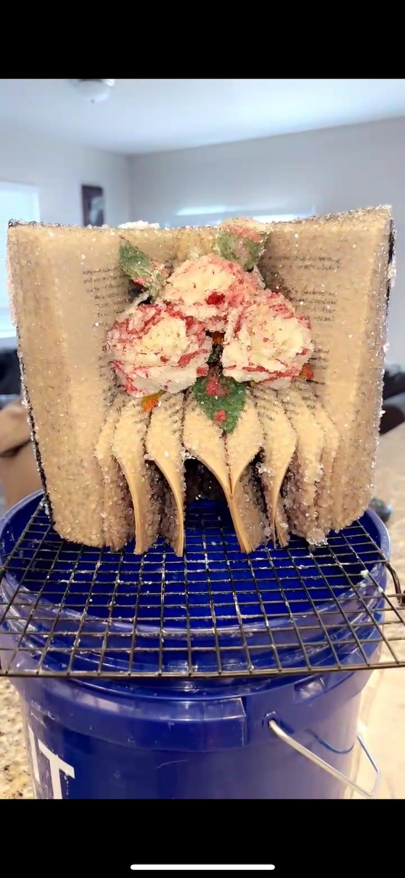May include: A unique art piece crafted from a book, shaped into a floral arrangement. The pages are visible, with a dusting of glitter. Red and white flowers with green accents adorn the top. The piece sits on a blue bucket with a metal grate.