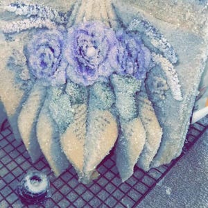 May include: A decorative, frosted cake with three lavender-colored rose accents and leaf details. The cake is on a wire rack, with two small, decorative elements nearby. The overall design is elegant and floral.