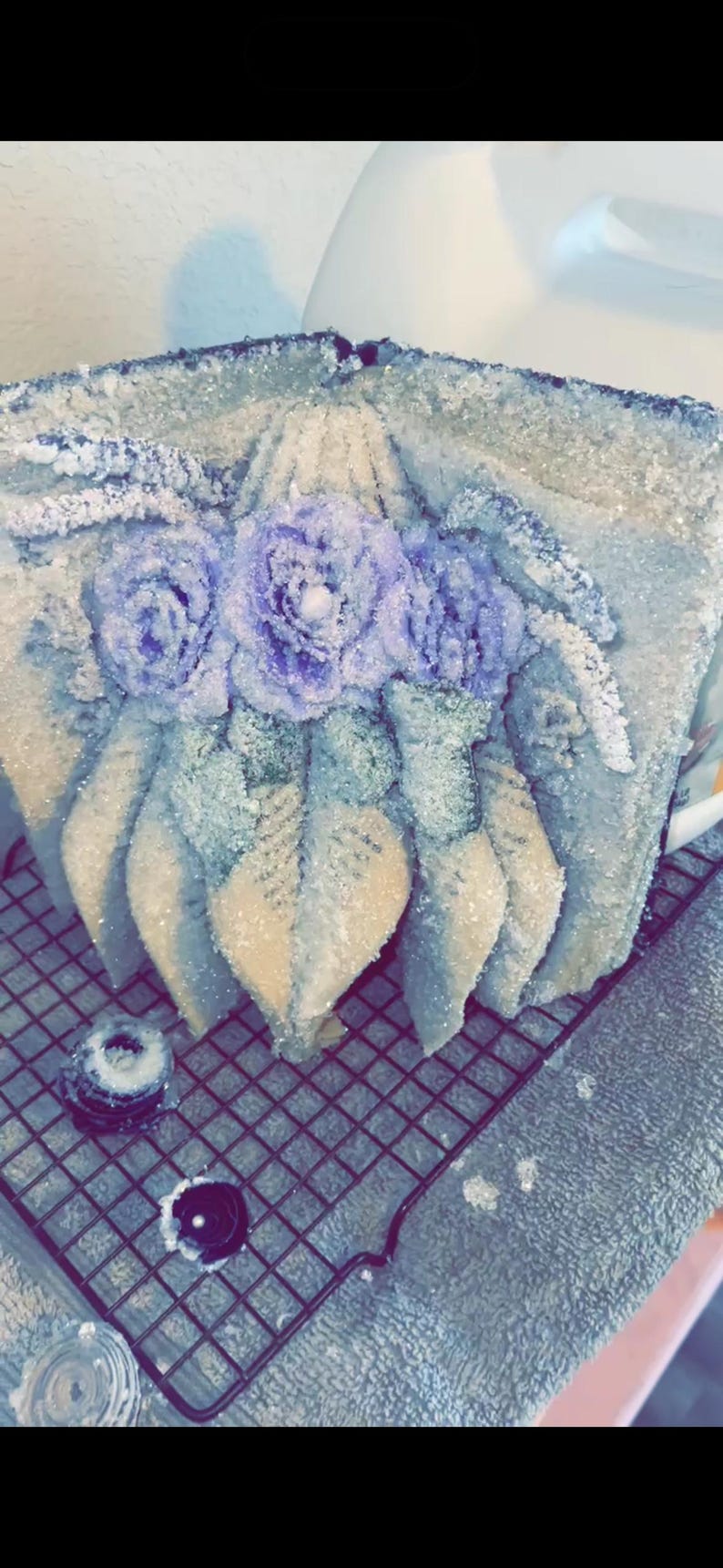 May include: A decorative, frosted cake with three lavender-colored rose accents and leaf details. The cake is on a wire rack, with two small, decorative elements nearby. The overall design is elegant and floral.