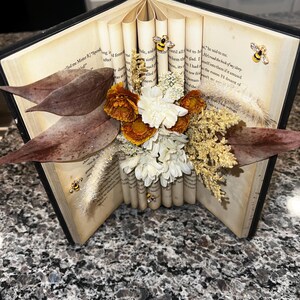 May include: An open book with pages rolled and adorned with dried flowers, leaves, and bee-shaped embellishments. The arrangement features orange, white, and brown floral elements, creating a unique decorative piece.