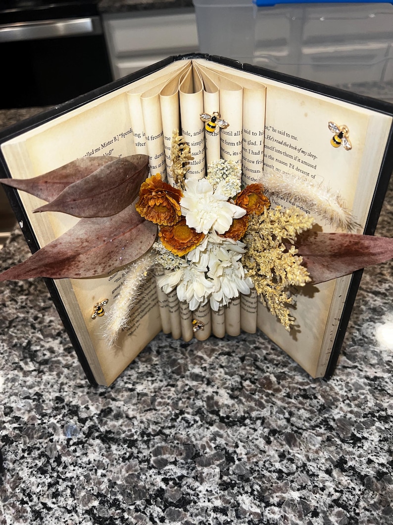 May include: An open book with pages rolled and adorned with dried flowers, leaves, and bee-shaped embellishments. The arrangement features orange, white, and brown floral elements, creating a unique decorative piece.