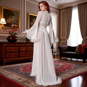 May include: A long, flowing white sheer robe with bell sleeves and ruffled trim. The robe features a high neckline and a cinched waist. The garment is worn in a room with a patterned rug and antique furniture.