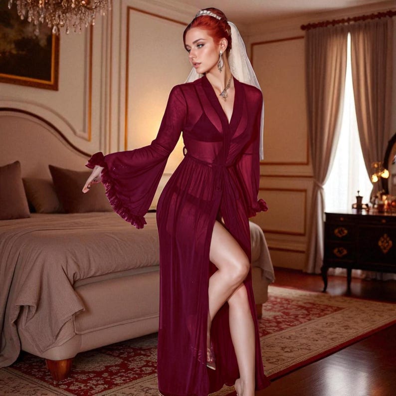 May include: A sheer burgundy robe with long, flared sleeves and a high slit. The robe is worn over a matching lingerie set. The model is wearing a veil and jewelry. The setting is a bedroom with a bed and a chandelier.