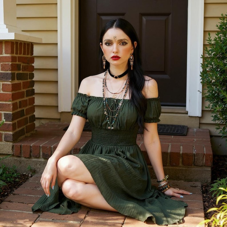 May include: An olive green off-the-shoulder dress with short sleeves. The dress has a fitted bodice and a flowing skirt. The person is wearing multiple necklaces, earrings, and bracelets. The person is sitting on a brick porch.