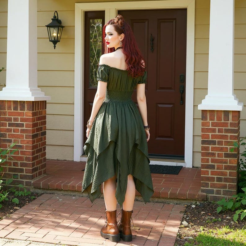 May include: An olive green off-the-shoulder dress with a tiered, asymmetrical hemline. The dress features short sleeves and a fitted bodice. The person is wearing brown boots. The background includes a brick and white column, a brown door, and a red brick walkway.