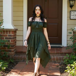 May include: An olive green off-the-shoulder dress with short sleeves and a layered skirt. The dress features a fitted bodice and a flowing, asymmetrical hemline. The model is wearing jewelry and boots.