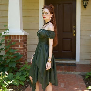 May include: An olive green off-the-shoulder dress with a layered, asymmetrical hemline. The dress features a fitted bodice and short sleeves. The model is wearing multiple bracelets, necklaces, and earrings. The background includes a brick and wood exterior.