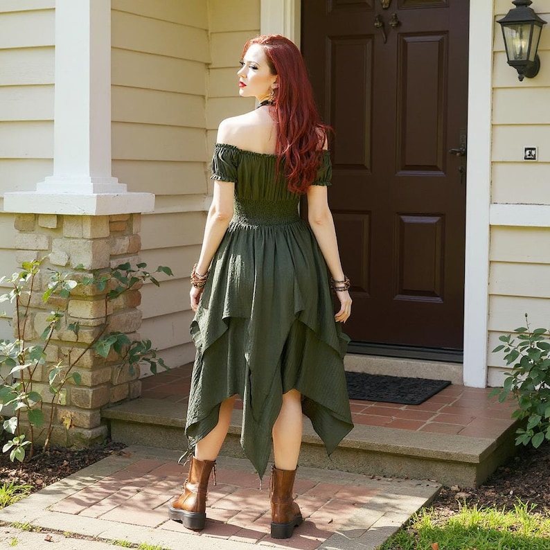 May include: An olive green off-the-shoulder dress with a tiered, asymmetrical hemline. The dress features a fitted bodice and short sleeves. The model is wearing brown boots. The background includes a house with a brown door and a brick walkway.