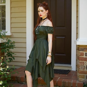 May include: An olive green off-the-shoulder dress with short sleeves and a layered, asymmetrical skirt. The dress has a fitted bodice and is worn by a person standing in front of a brown door. The person is wearing jewelry, including a necklace and bracelets.