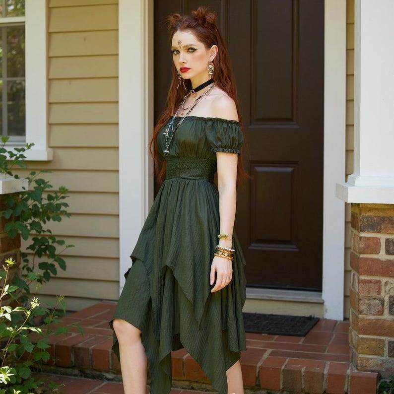 May include: An olive green off-the-shoulder dress with short sleeves and a layered, asymmetrical skirt. The dress has a fitted bodice and is worn by a person standing in front of a brown door. The person is wearing jewelry, including a necklace and bracelets.