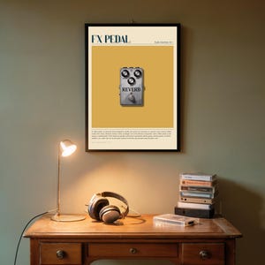 May include: A framed poster with the text "FX PEDAL" and a silver reverb pedal illustration. The poster is centered on a mustard yellow background. A desk lamp, headphones, and books are on a wooden desk below the poster.