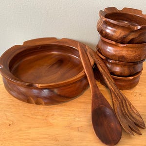 May include: A set of wooden salad bowls and serving utensils. The set includes a large round bowl, a stack of smaller bowls, a spoon, and a fork. The bowls and utensils are made of a dark brown wood.