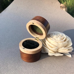 May include: A small, cylindrical wooden ring box featuring a dark brown exterior with a lighter wood trim. The box has a hinged lid, a black velvet interior lining, and a small magnetic closure. Beside the box sits a white wooden flower.