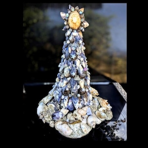 May include: A conical decorative sculpture constructed from an assortment of seashells in shades of blue, white, and tan. The structure features a pointed top adorned with a single, larger, light-coloured shell, with smaller shells layered below.