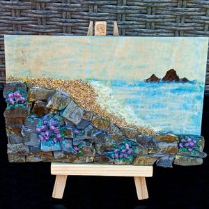 May include: A mixed-media artwork showing a coastal scene. It features a stone wall with purple flowers, a sandy beach, and a blue ocean with two small islands. The artwork is displayed on a small wooden easel.
