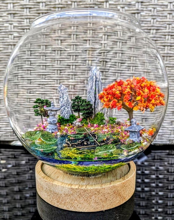 A hand-assembled miniature diorama lamp with a brass boatman, lily pads, and cherry blossom tree that casts a serene, layered glow — it's practically a tiny world you plug in.