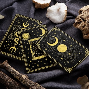 Könnte beinhalten: Drei schwarze Tarotkarten mit goldenen Himmelsdesigns, darunter Monde, Sonnen und Sterne. Die Karten sind auf einem dunkelgrauen Stoff angeordnet, mit dekorativen Rändern und komplizierten Illustrationen. Das Gesamtthema ist mystisch und esoterisch.