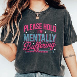 May include: Dark gray t-shirt with the text "PLEASE HOLD I'M MENTALLY BUFFERING" in pink and blue. The shirt has a vintage, worn look. The design includes stars and a loading bar graphic.