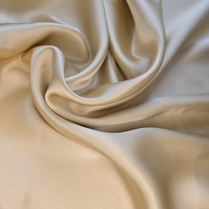Può includere: Un pezzo di tessuto color champagne con una superficie liscia e lucente e un drappeggio morbido e fluido. Il materiale presenta sottili riflessi e ombre profonde create dalle pieghe, mostrando una consistenza uniforme, riflettente e satinata.