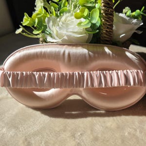 Puede incluir: Un antifaz para dormir de color rosa pálido con un acabado suave similar al satén y una banda elástica fruncida a juego. El antifaz presenta una forma contorneada diseñada para cubrir los ojos, con una construcción acolchada y suave.