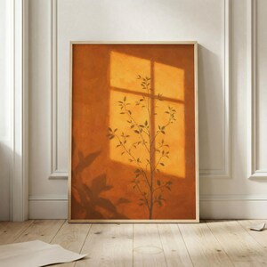 May include: A rectangular wall art print in a light wood frame featuring a warm, burnt orange background. The image displays the silhouette of a slender plant with small leaves, cast by a window grid shadow that creates a geometric pattern.