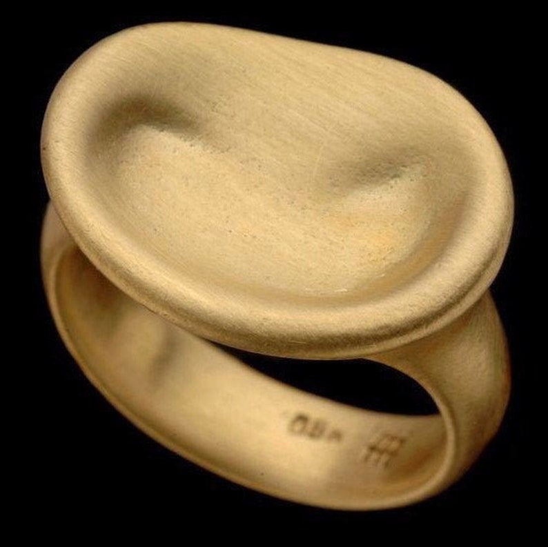 Puede incluir: Un anillo de oro con una parte superior lisa, redondeada y ligeramente c&oacute;ncava. El anillo est&aacute; hecho de metal cepillado y tiene un dise&ntilde;o simple y moderno.