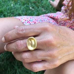 Puede incluir: Un anillo de oro con una parte superior grande de forma ovalada. La parte superior del anillo es lisa y tiene una forma ligeramente c&oacute;ncava.