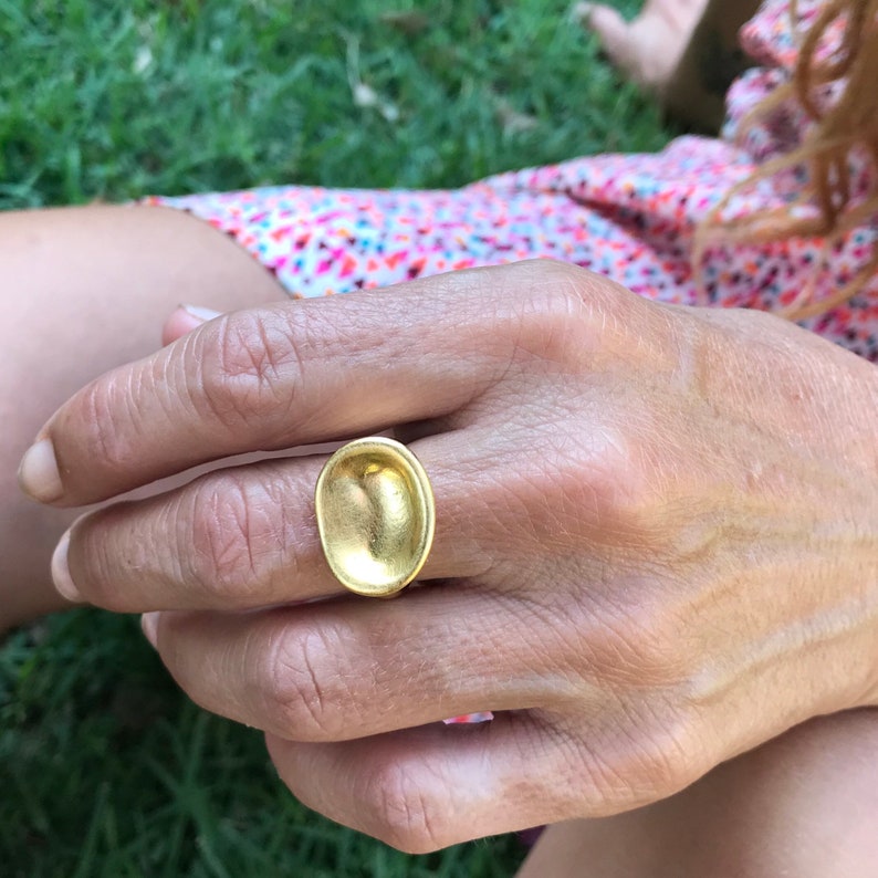 Puede incluir: Un anillo de oro con una parte superior grande de forma ovalada. La parte superior del anillo es lisa y tiene una forma ligeramente c&oacute;ncava.