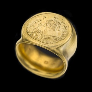 May include: A gold ring with a raised design of a crowned figure on the top. The ring is made of a smooth, polished metal.