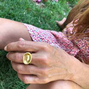 Puede incluir: Un anillo de oro con una parte superior grande de forma ovalada. El anillo se lleva en un dedo.