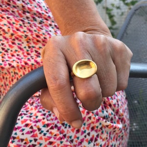 Puede incluir: Un anillo de oro con una parte superior grande de forma ovalada. El anillo se lleva en un dedo.