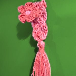 Puede incluir: Un llavero rosa hecho a mano con ganchillo, con una flor, cuerpo trenzado y borla. El llavero está sujeto a un anillo de color plateado. Los colores del hilo van del rosa claro al rosa oscuro.
