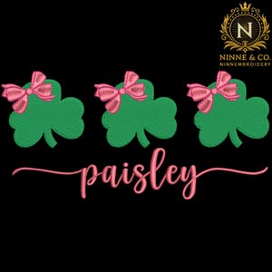 May include: Embroidered design featuring three green shamrocks, each adorned with a pink bow. The cursive word "paisley" is embroidered in pink below the shamrocks. The background is black.