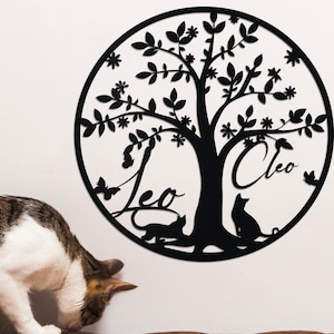 Puede incluir: Un adorno de pared de metal negro con un diseño de árbol dentro de un marco circular. El árbol tiene hojas, flores y los nombres "Leo" y "Cleo" con siluetas de gatos. Un gato atigrado está en un sofá de cuero marrón.