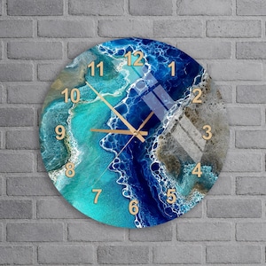 May include: A round wall clock featuring an abstract ocean-inspired design with swirling shades of deep blue, turquoise, and sandy beige. The clock face displays gold-coloured numerals from 1 to 12 and matching gold-coloured hour, minute, and second hands.