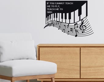 Puede incluir: Arte de pared de metal negro con un teclado de piano, notas musicales y el texto "IF YOU CANNOT TEACH ME TO FLY, TEACH ME TO SING". La obra de arte está montada en una pared blanca sobre un mueble de madera.